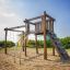 Kampinos wooden playhouse with slides and climbing net on a sandy playground.