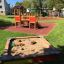 Sandbox with toy trucks and playground equipment in the background on a sunny day.