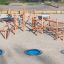 Sonora wooden playground climbing structure with blue trampolines on sandy ground.