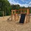 Alstad wooden playground equipment with slide, climbing wall, and swings in a park setting.