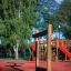 Wooden Barrel playground equipment on a red safety surface with trees and playground in the backgrou