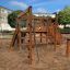Kiosk Modern Nature wooden playhouse on a sandy playground surrounded by buildings and trees.